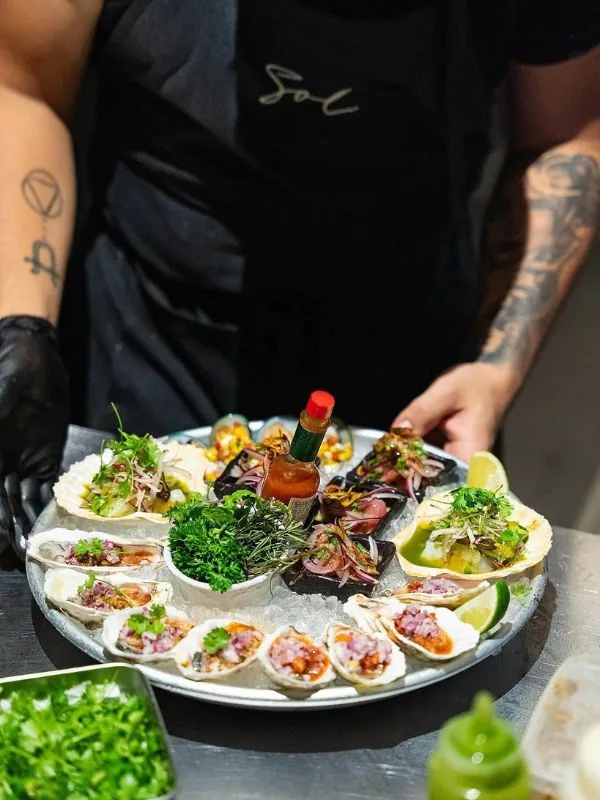 Nhân viên SOL bưng đĩa hải sản hàu tươi trên vỏ với hành tím, rau thơm và sốt salsa, kèm chanh và sốt cay, phong cách Mỹ Latin